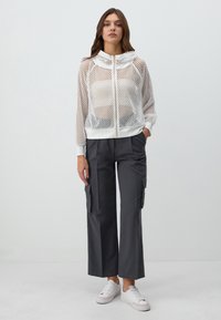 Giacca bianca a rete con zip e cappuccio, abbinata a pantaloni cargo grigi scuri a gamba larga con tasche laterali e sneakers bianche.