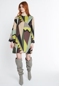 Robe à motifs avec manches longues, présentant des formes géométriques en vert, beige, marron et noir. Associée à des bottes en tissu grises jusqu'au genou.