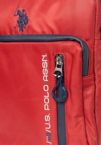 Červený batoh s tmavě modrým zipem, vyšitým a potištěným logem U.S. Polo Assn., s kruhovým jezdcem na zipu s textem značky.