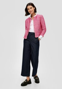 Vaaleanpunainen tweed-takki, jossa on pyöreä kaula-aukko, yhdistettynä tummiin leveisiin housuihin ja mustiin loafer-kengiin, joissa on kultaiset yksityiskohdat.