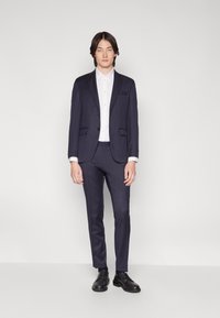 Navy tailored suit with two-button jacket, notch lapels, and slim-fit trousers. White button-up shirt beneath, paired with black dress shoes.