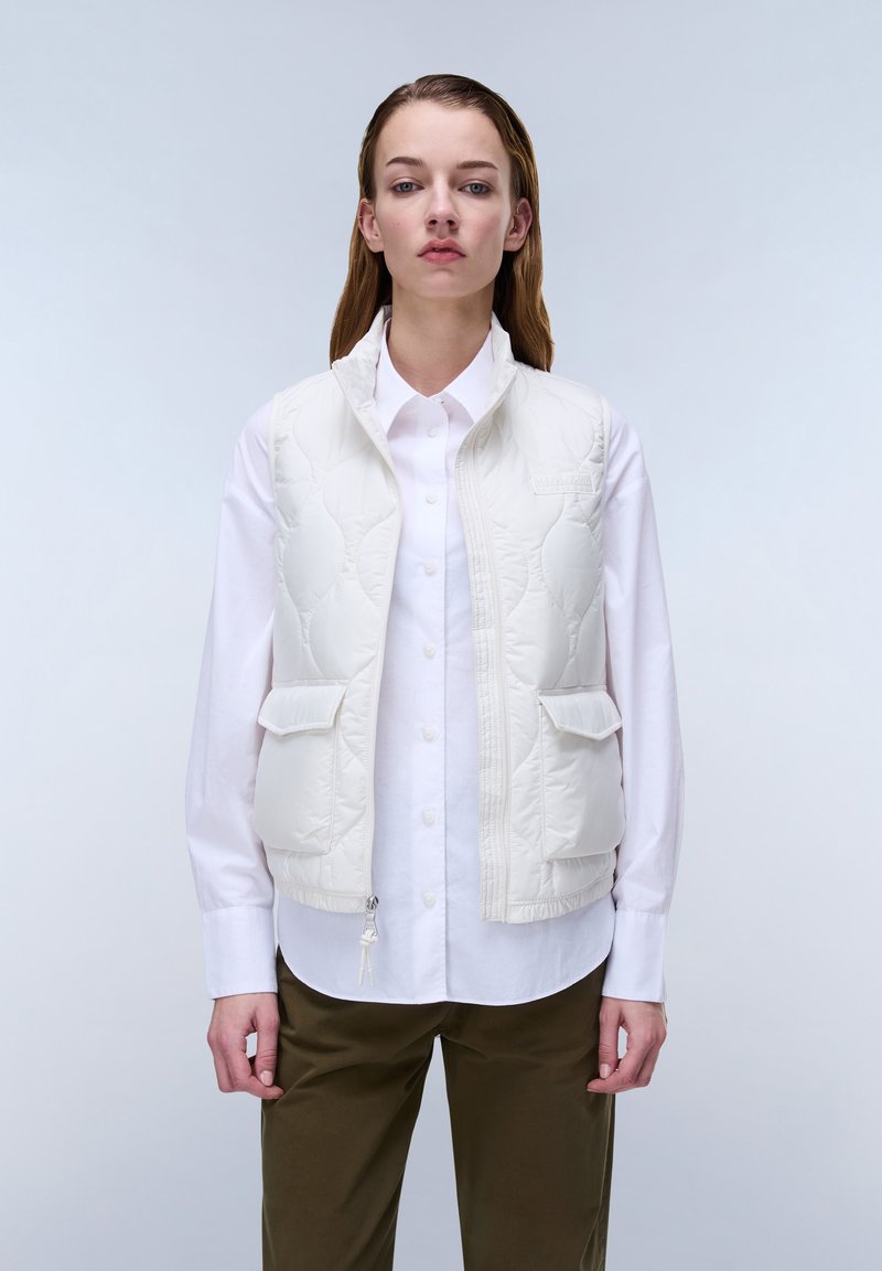 Mujer con chaleco acolchado blanco sobre una camisa blanca abotonada y pantalones verde oliva, de pie frente a un fondo claro y liso.