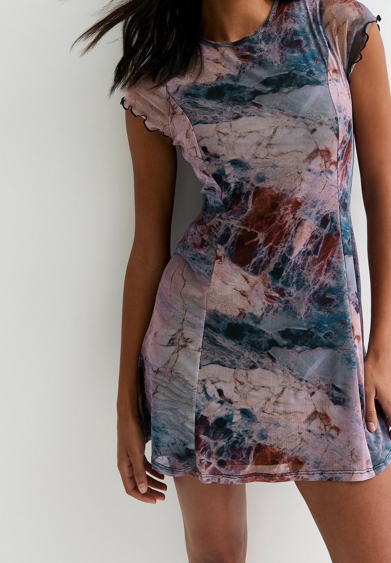 Femme portant une robe courte ajustée avec un motif abstrait en marbre multicolore et des manches courtes à volants sur un fond clair uni.