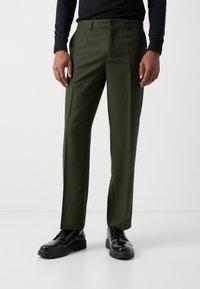 Pantalons ajustés verts avec une coupe slim, dotés d'une fermeture à bouton et d'un devant plat. Associés à des chaussures noires à lacets pour créer un contraste.