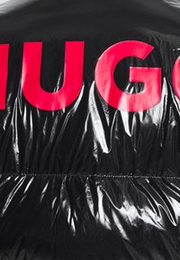Černá, lesklá péřová bunda s velkým růžovým logem "HUGO" na hrudi. Texturovaný povrch s nařaseným švem v pase.