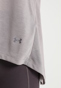 Camiseta deportiva de color gris claro hecha de una tela suave y liviana. Cuenta con un dobladillo redondeado, un logo de marca prominente cerca de la parte inferior y mangas cortas.