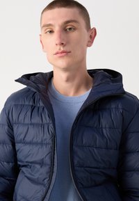 Jūras spēku zils pūkainais mētelis ar horizontālu kanālu kviltu, ar kapuci un ar rāvējslēdzēju priekšpusē. Valkāts virs gaiši zila džempera. Mīksta, gluda faktūra.