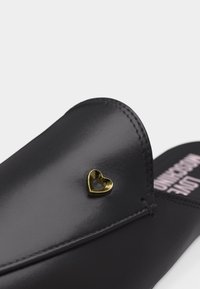 Μαύρη δερμάτινη μοκασίνι με ένα μικρό χρυσό μεταλλικό διακοσμητικό σε σχήμα καρδιάς στο πάνω μέρος κοντά στα δάχτυλα.