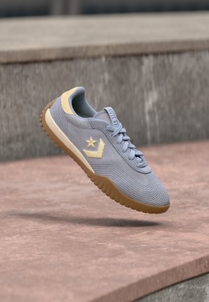 Pantof sport albastru deschis cu siglă bej și talpă gumă plutind deasupra unor trepte din piatră într-un mediu urban în aer liber.