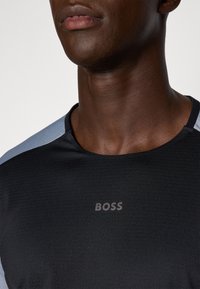 Svart treningstrøye med lysegrå detaljer, laget av teksturert stoff og med en subtil "BOSS"-logo på brystet. Rund hals, tettsittende design.