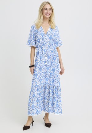 Naeratav naine, kes kannab sinise ja valge mustriga lühikeste varrukatega maksikleit ning musti kontsakingi, seisab ühevärvilise tausta ees.