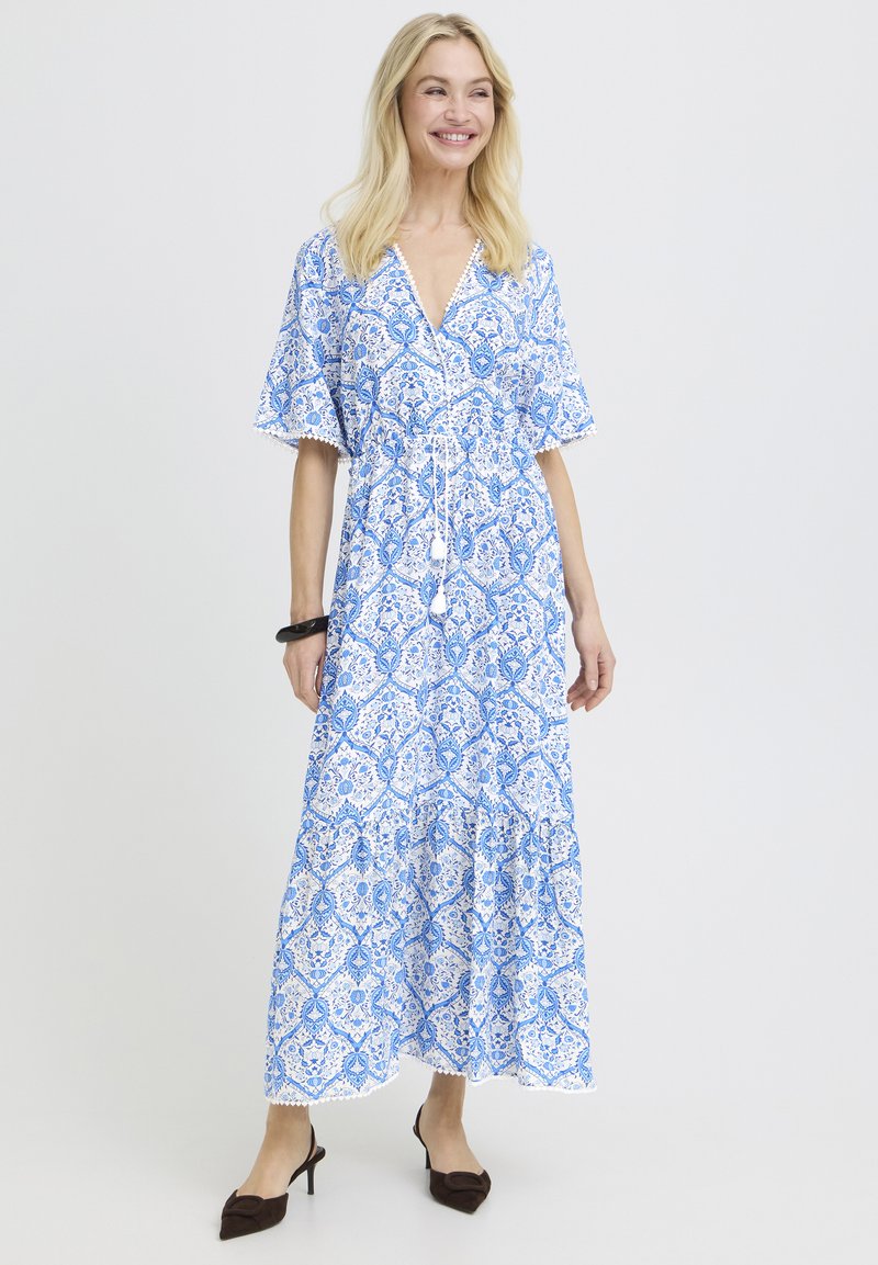 Femme souriante portant une robe longue à motifs bleus et blancs avec des manches courtes et des chaussures à talons noires, debout devant un fond uni.