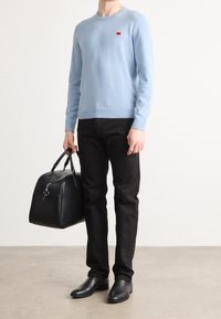 Maglione a maglia azzurro chiaro con un piccolo logo rosso, jeans neri, borsa in pelle nera con doppio manico e stivali alla caviglia neri. Design minimalista.
