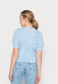 Blusa azul com padrão floral, mangas bufantes, cintura franzida e detalhe de buraco na parte de trás. Tecido leve com pequenos padrões de flores brancas e azuis.