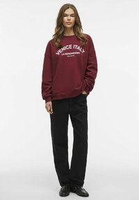 Felpa bordeaux con testo bianco che dice "Venezia Italia" e "La Serenissima", abbinata a pantaloni neri e scarpe nere.
