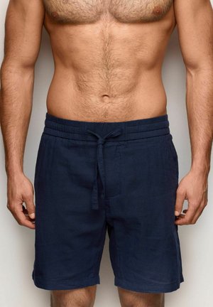 Mężczyzna bez koszulki z lekką opalenizną, ubrany w granatowe szorty z troczkami, stoi z luźno opuszczonymi ramionami przy ciele na neutralnym tle.