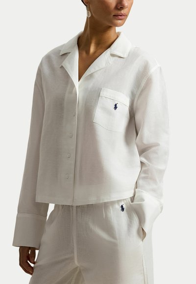 Vrouw draagt een witte blouse met lange mouwen en bijpassende broek met geborduurde marineblauwe logo's op de zak en heup.