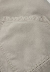 Gros plan sur une poche en tissu beige avec un tissage texturé visible et des coutures doubles sur un vêtement.