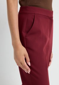 Bordeaux kleurige broek met een soepele textuur, voorzien van een zijzak en plooien, die een op maat gemaakt ontwerp benadrukken. Hand rustend op de tailleband.