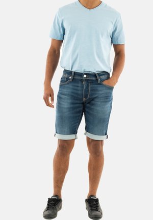 Chemisette bleu clair à manches courtes et décolleté en V, associée à un short en denim bleu foncé retroussé, portée avec des baskets noires.
