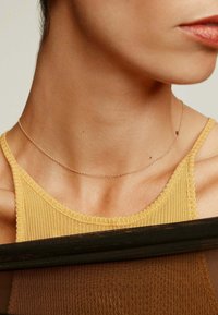 Un delicado collar de oro reposa sobre un cuello desnudo, con finos eslabones visibles. El fondo es una tela oscura y texturizada que contrasta con un tono de piel claro.