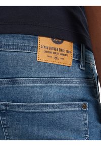 Jeans de mezclilla azul con una etiqueta de cuero marrón, con detalles de costura, bolsillos traseros y etiqueta de marca. Diseño casual con una forma ajustada.