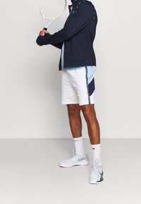 Marinblå jacka med vita accenter, ljusblå t-shirt, vita shorts med marinblå sidopanel, ihop med vita Nike-sneakers och en tennisracket.