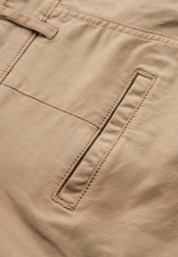 Beige stoflomme med lodret åbningslomme og synlige syninger, del af et beklædningsstykke.