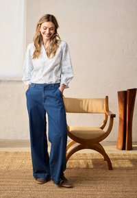 Femme souriante aux longs cheveux portant un chemisier blanc et un jean large bleu, debout à côté d'une chaise en bois dans une pièce lumineuse.