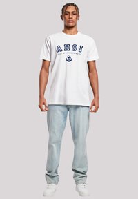 White cotton T-shirt with "AHOI" printed in navy blue, featuring an anchor graphic and relaxed fit. Paired with light blue denim jeans.