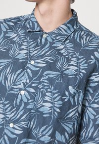 Camicia a maniche corte con bottoni in tessuto blu scuro, caratterizzata da un motivo foglioso in blu chiaro, con colletto e taschino sul petto.