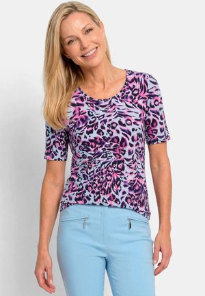 Vrouw glimlacht, draagt een paars-roze dierenprint shirt met korte mouwen en lichtblauwe broek met voorzakken met rits, staand tegen een effen achtergrond.