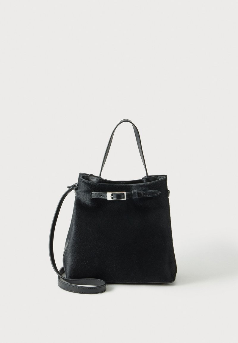 Sac à main en cuir noir avec poignée supérieure, sangle longue réglable et détail de boucle de ceinture à l’avant sur un fond clair uni.