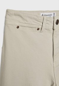Pantalon beige taille haute avec passants pour ceinture, rivets métalliques et une étiquette de taille blanche indiquant "Stradivarius 38 USA & MEX".