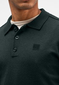 Dunkelgrünes Poloshirt mit strukturiertem Strick, drei Knöpfen am Kragen und einem kleinen schwarzen Logo-Patch auf der Brust.