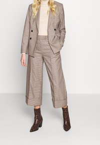 Kvinna som bär beige och brun rutiga vida cropped byxor tillsammans med en matchande dubbelknäppt blazer och bruna mönstrade ankelskor.