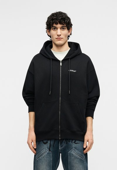 Νεαρός με κατσαρά μαλλιά, φοράει μαύρο φούτερ με φερμουάρ και κορδόνια, καθώς και μπλε τζιν με Ραφές, στέκεται μπροστά σε λευκό φόντο.