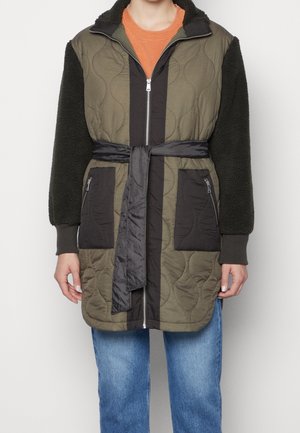 Persoon draagt een doorgestikte olijfgroene en zwarte jas met ceintuur in de taille, rits aan de voorkant, met rits afgesloten zakken, zwarte fleece mouwen, oranje shirt en blauwe spijkerbroek.