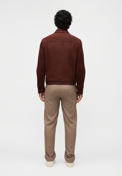 Άνδρας που στέκεται με την πλάτη, φοράει σκούρο καφέ μπουφάν, μπεζ παντελόνια και λευκά αθλητικά παπούτσια σε απλό ανοιχτό φόντο.