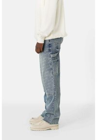 Hellblaue Jeans im geraden Schnitt, mit Seitentaschen und subtilen Abnutzungserscheinungen. Kombiniert mit einem weißen Langarmhemd und beigen Schuhen.