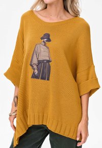 Żółty sweter musztardowy z szerokimi rękawami, prążkowanym dołem i przednim nadrukiem postaci w liniowych wzorach. Miękka tekstura, luźny fason.