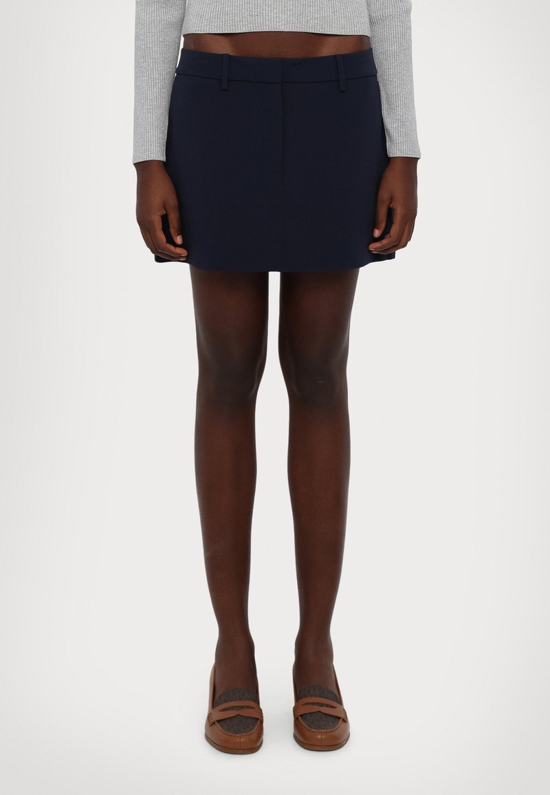 Mini-jupe bleu marine à coupe ajustée, en tissu lisse. Elle présente une ceinture plate et des poches latérales. Portée avec des mocassins marron.