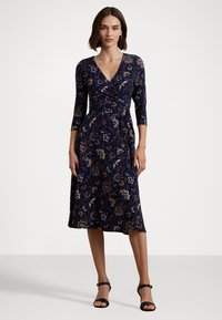 Abito avvolgente in tessuto blu scuro con motivi floreali e paisley, maniche a tre quarti e lunghezza midi. Abbinato a sandali neri con cinturini.