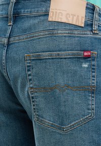 Gaiši zili džinsi ar aizmugurējo kabatu, kurā redzams oranžs zigzag šuvju raksts. Satur brūnu ādas birku ar uzspiestu uzrakstu "BIG STAR".