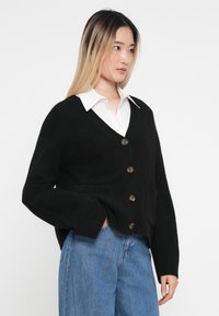 Svart stickad cardigan med V-hals, fem bruna knappar och två framfickor, bärs över en vit skjorta med krage och blå jeans.