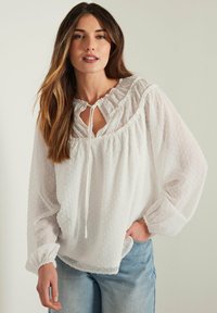 Blusa bianca con pois testurizzati, scollo a volant e dettaglio con cordino. Maniche lunghe con polsini elasticizzati, tessuto leggero e trasparente.