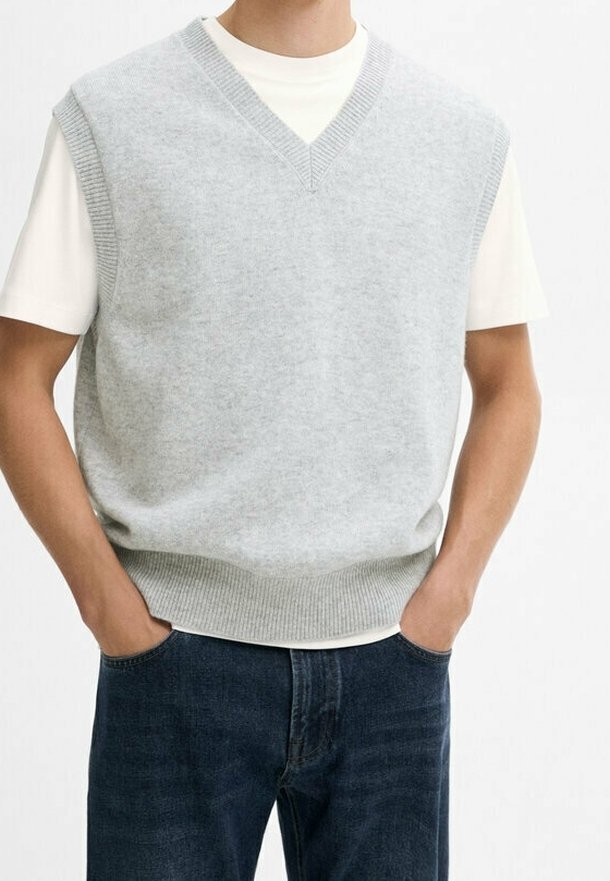 Personne portant un gilet en maille gris clair à col en V par-dessus un T-shirt blanc à manches courtes avec un jean bleu foncé, les mains dans les poches.