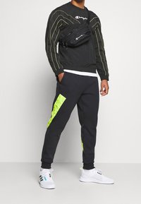 Felpa sportiva nera con accenti giallo neon, abbinata a pantaloni della tuta neri con un pannello laterale giallo e calzature bianche.