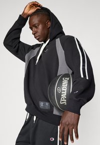 Sweatshirt preto com detalhes cinzas e riscas brancas, em material de algodão. O modelo segura uma bola de basquete Spalding preta, vestindo calções pretos a condizer.