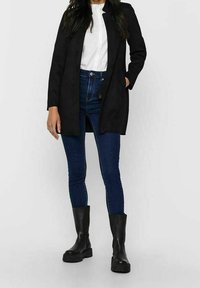 Blazer nero a maniche lunghe con un design elegante, texture morbida e davanti aperto. Abbinato a jeans blu aderenti e stivali neri alla caviglia.